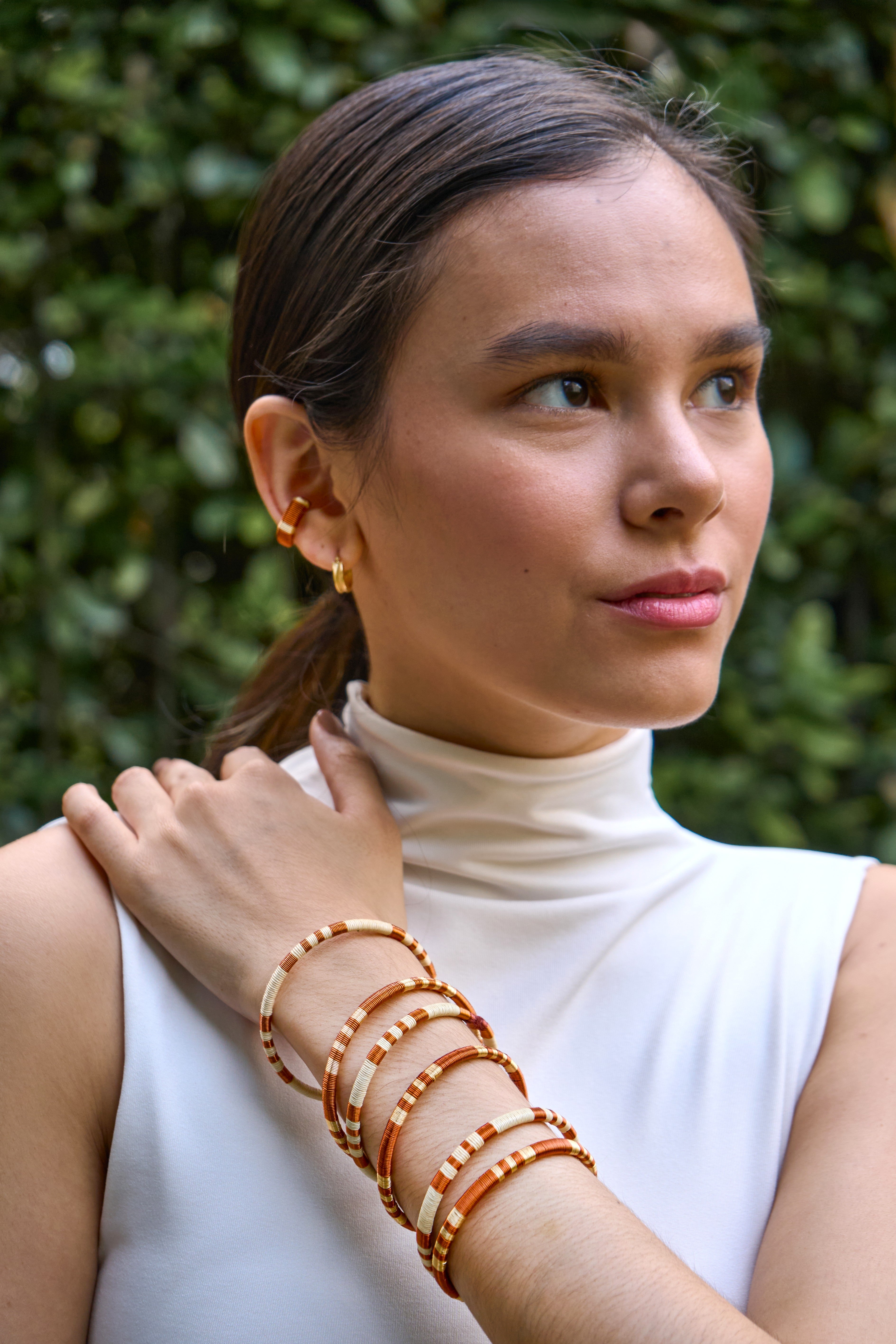 Woman wearing gold copper ear cuffs and bracelets against a blurred green background