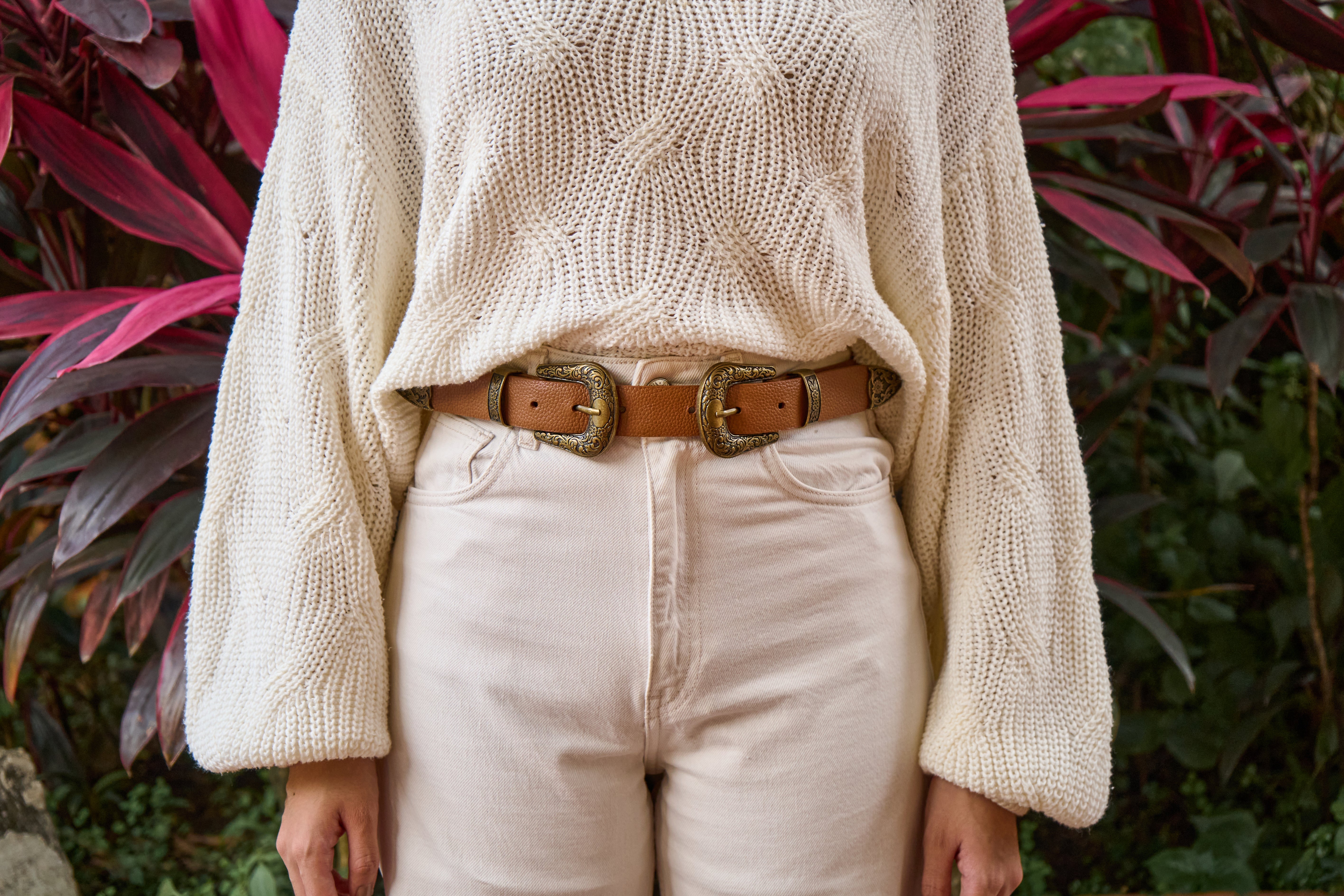 Person wearing a beige sweater and light-colored pants with a brown belt, standing against a natural background.